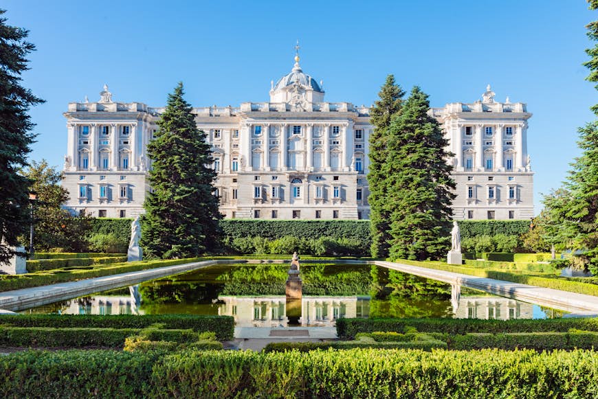 A view of the grand, white Royal Palace in Madrid, viewed from the accompanying gardens.  The historic-looking building is reflected in a small pond, which is also located on the grounds.