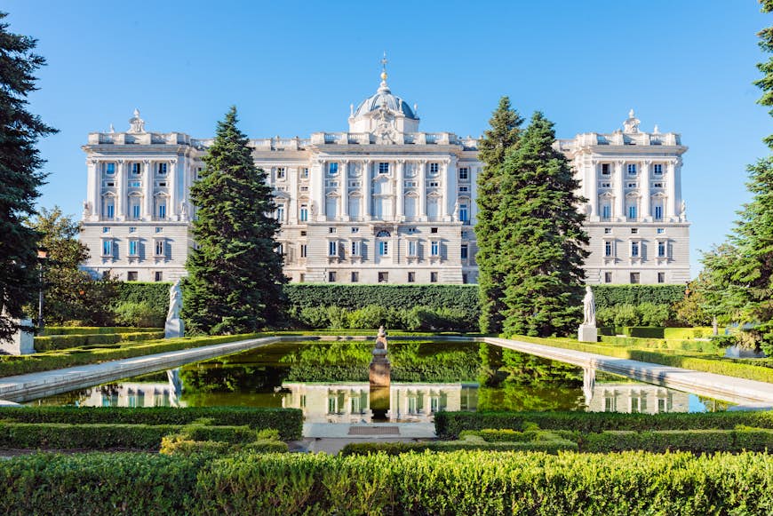 A view of the grand, white Royal Palace in Madrid, viewed from the accompanying gardens. The historic-looking building is reflected in a small pond, which is also located on the grounds.