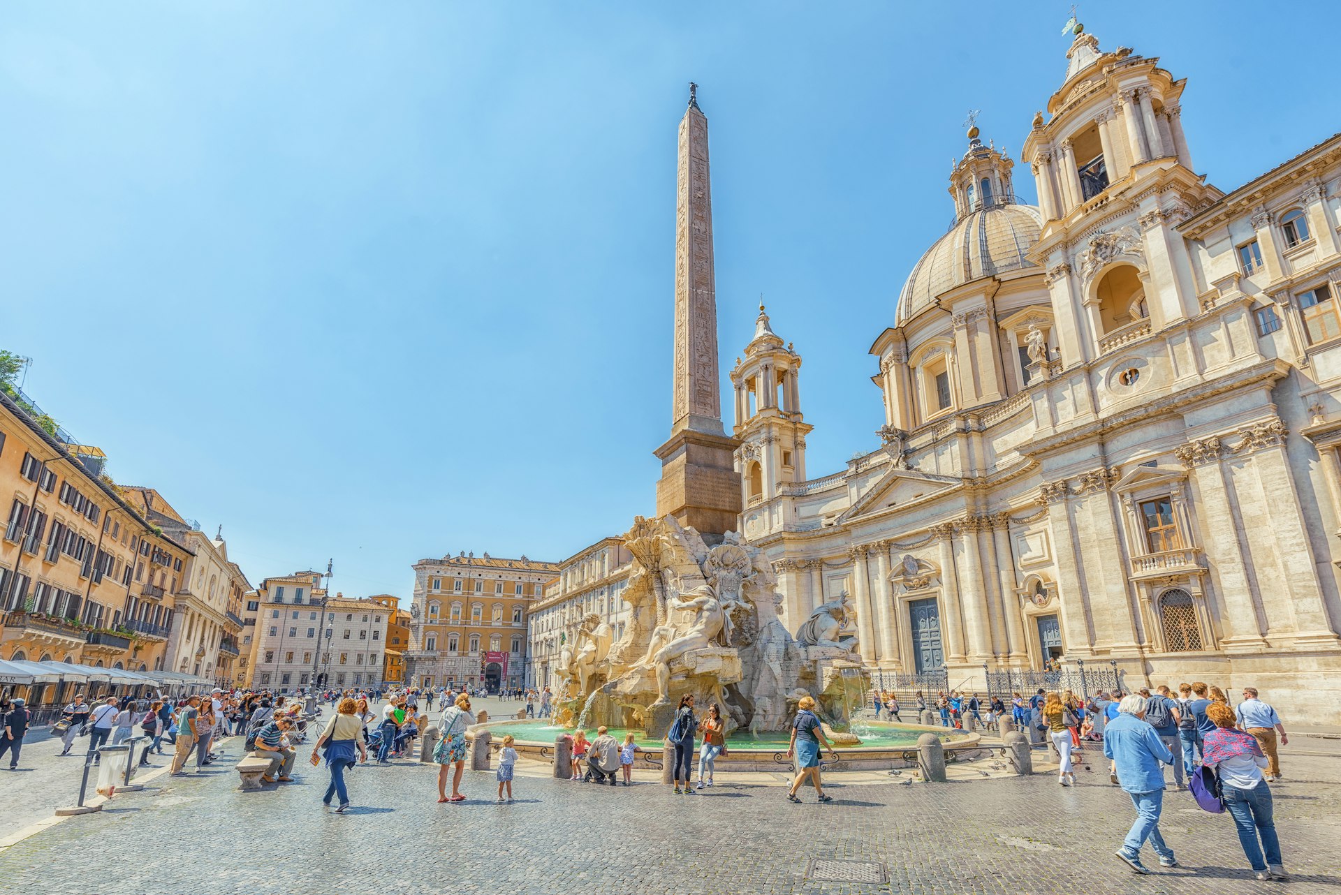 A square lined with grand buildings. A freestanding obelisk surrounded by fountains is the centerpiece