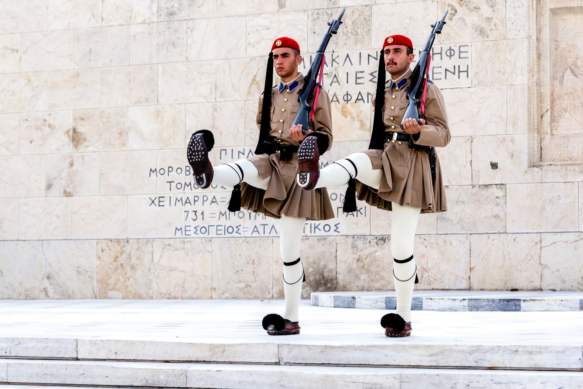Two guards in front of the rose-toned Hellenic Parliament building in Syntagma Square, Athens, march in time during the changing of the guard. Both are dressed in traditional evzones costume, a tasseled fez hat, thick kilt and stockinged legs ending at pom-pom shoes.