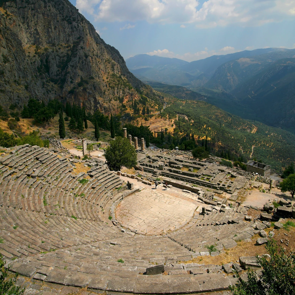 A view from the top row seats at the Theatre of Delphi, Greece. Beyond the circular theatre, vast forested valleys are visible.
