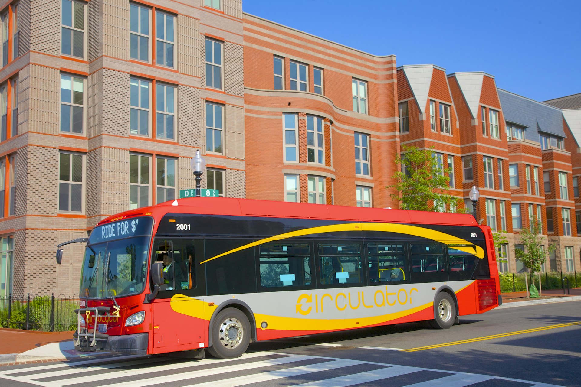 A bright red and yellow DC Circulator bus drives past a tan and orange brick building in Washington, DC