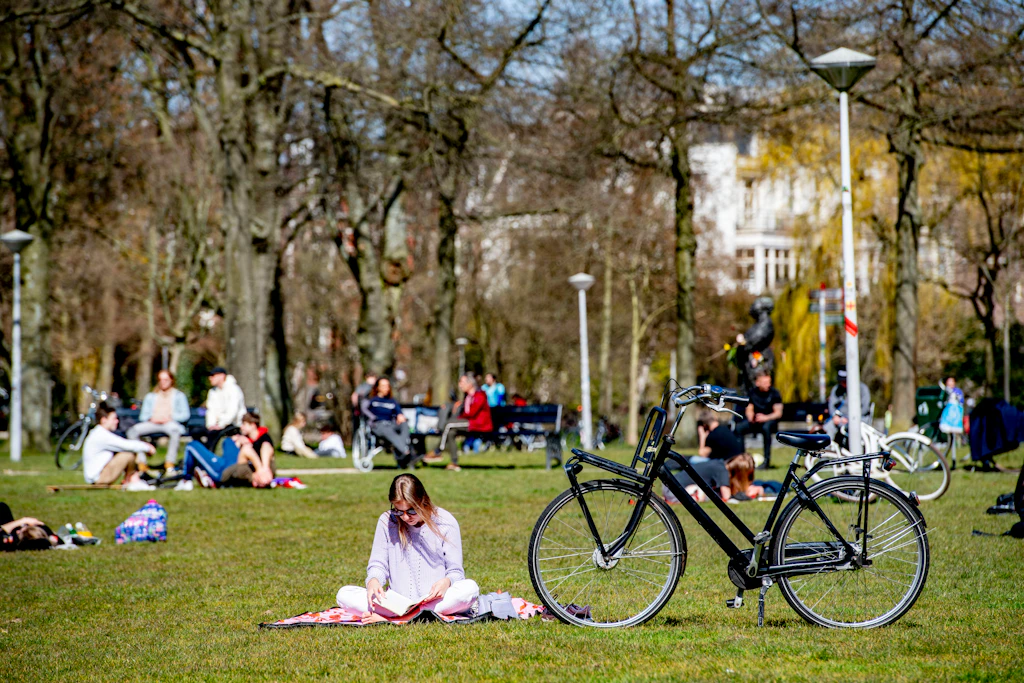 A woman sits cross legged and reads a book in the sunshine during a spring day at the Vondelpark in Amsterdam. She is sat next to her black bicycle with trees and other park users in the background.