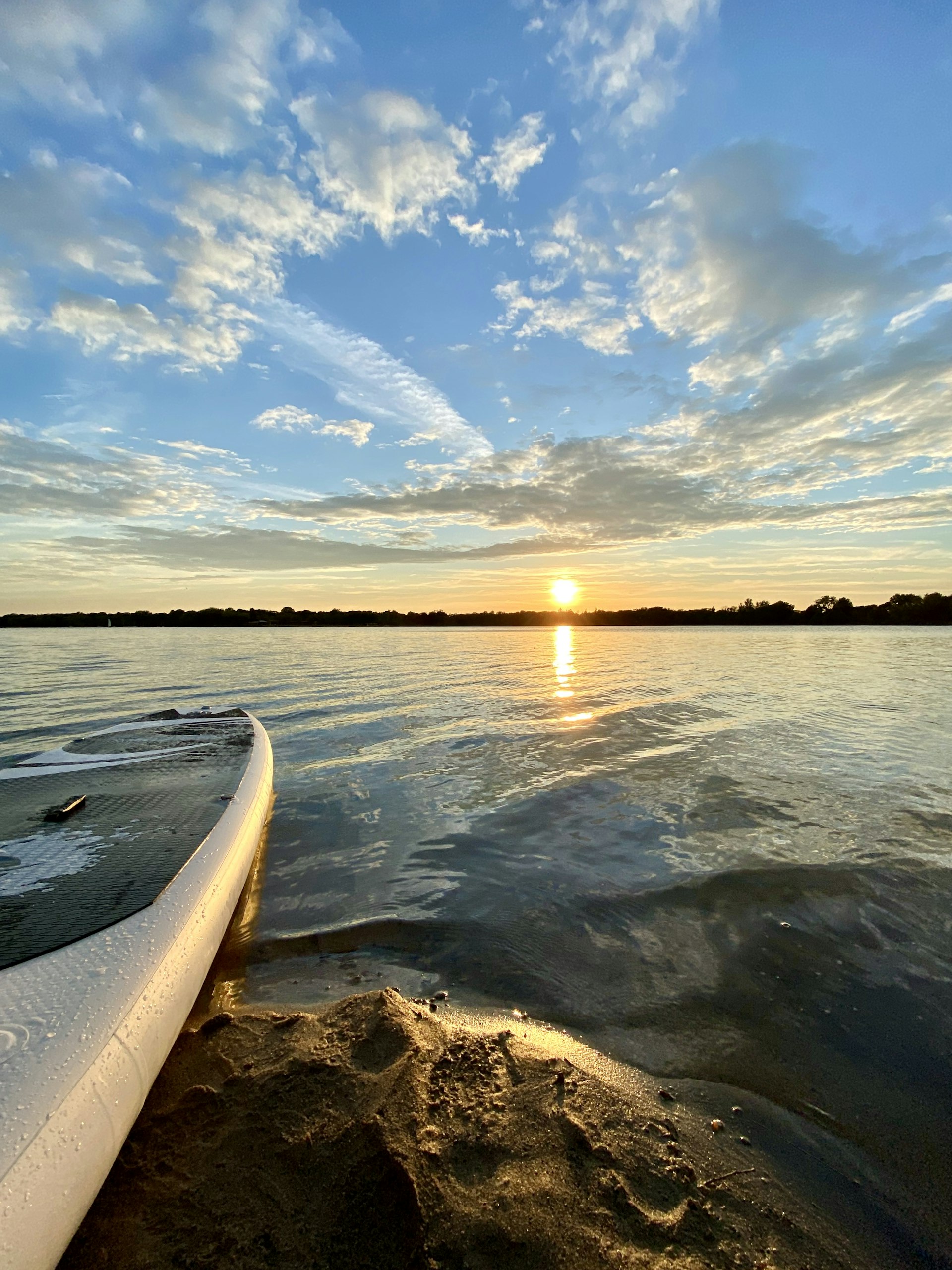 A shot looking down a paddleboard out onto a lake as the sun sends orange streaks across a blue cloudy sky