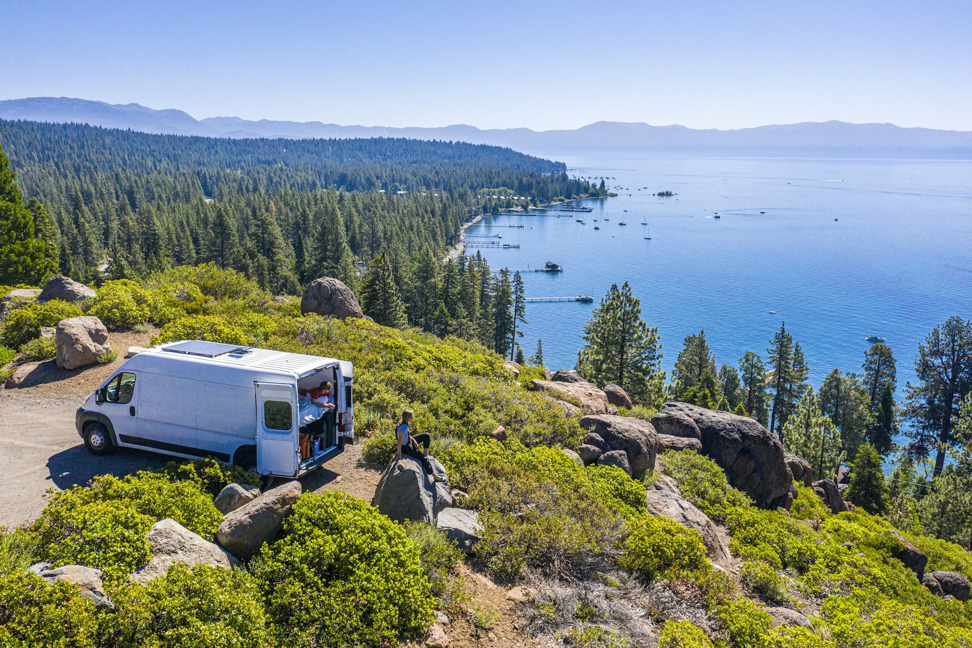 Aerial shot of a campervan at a viewpoint with two people sat overlooking a lake