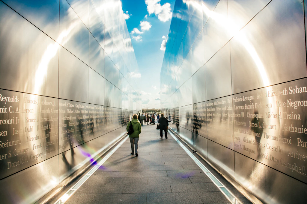 People in silhouette walk through a passage between large plinths with names engraved on them. Light reflects off the walls creating a circle of sunlight