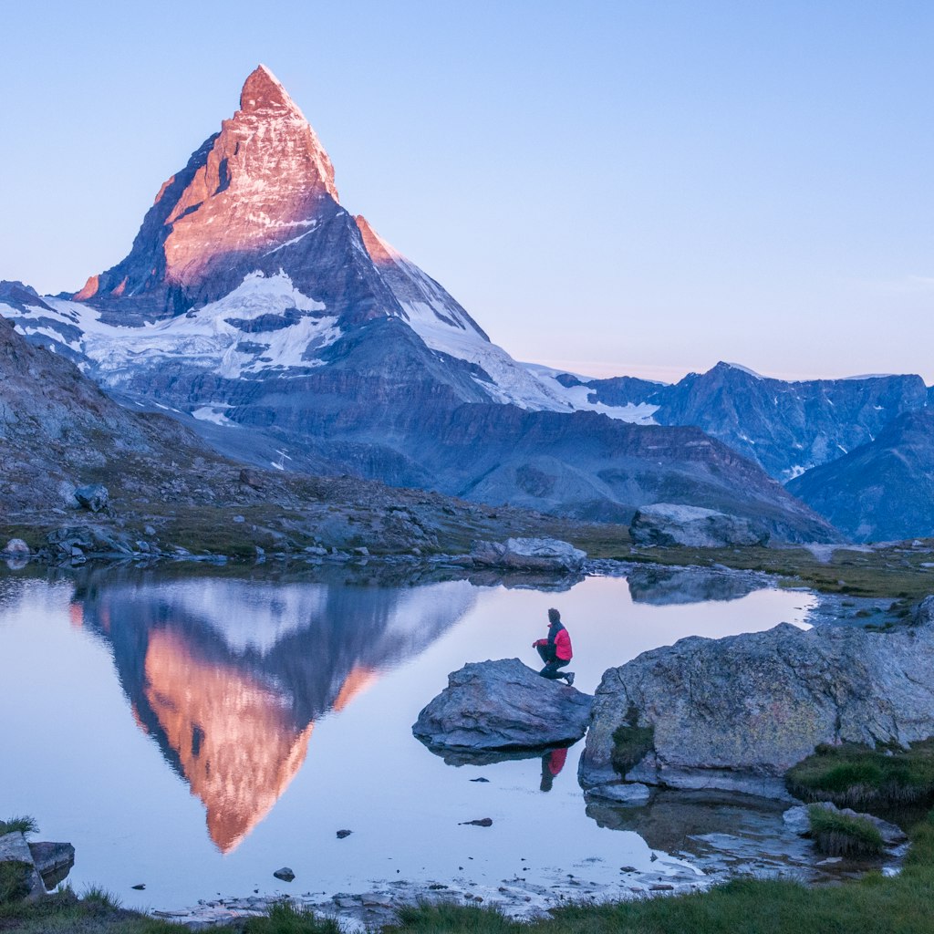 Early morning dawn scene of sunrise on the Matterhorn Mountain reflecting pink in the lake with male man on rock with red down jacket with clear blue sky Gornergrat Zermatt Matterhorn Europe