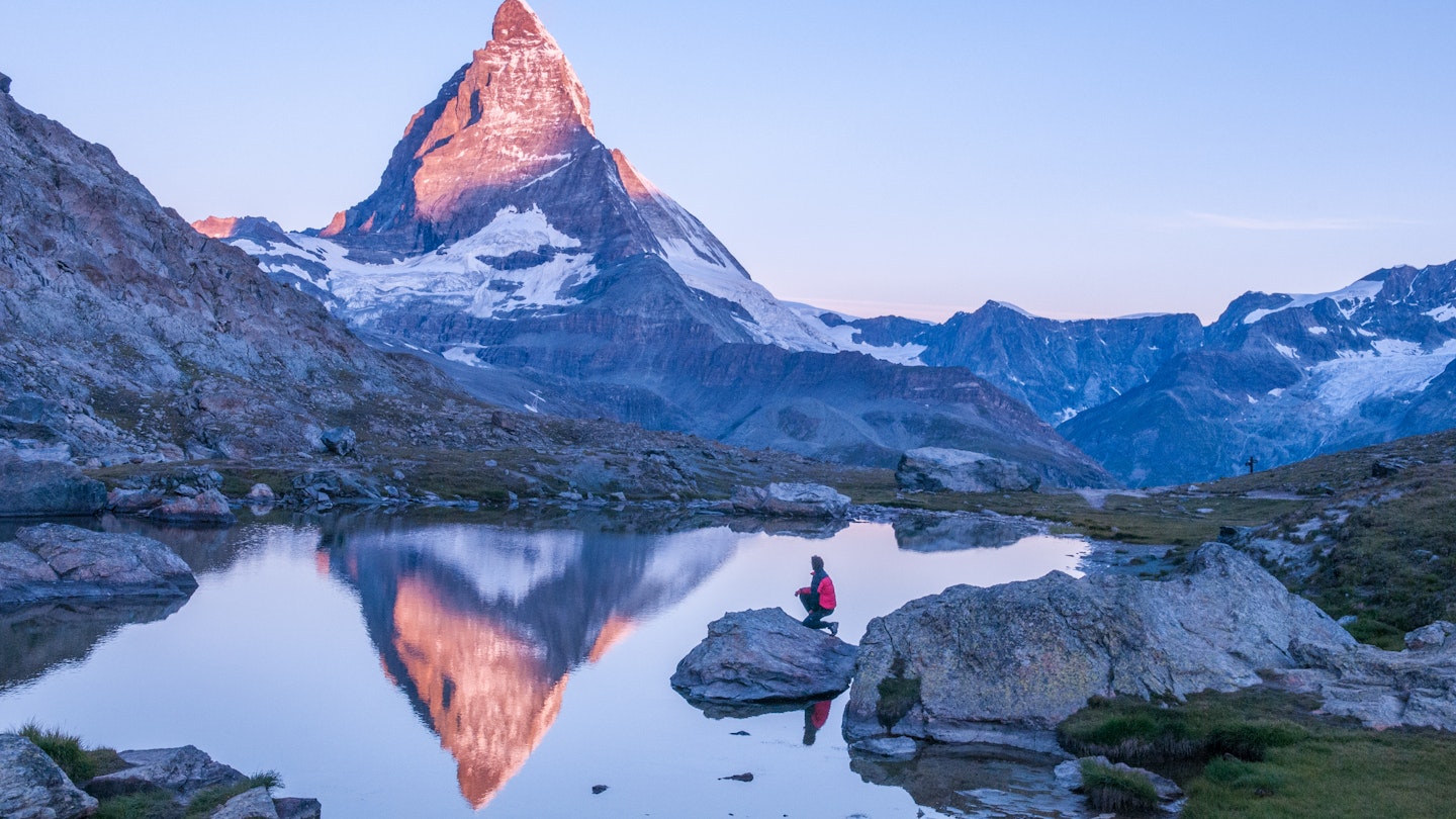 Early morning dawn scene of sunrise on the Matterhorn Mountain reflecting pink in the lake with male man on rock with red down jacket with clear blue sky Gornergrat Zermatt Matterhorn Europe