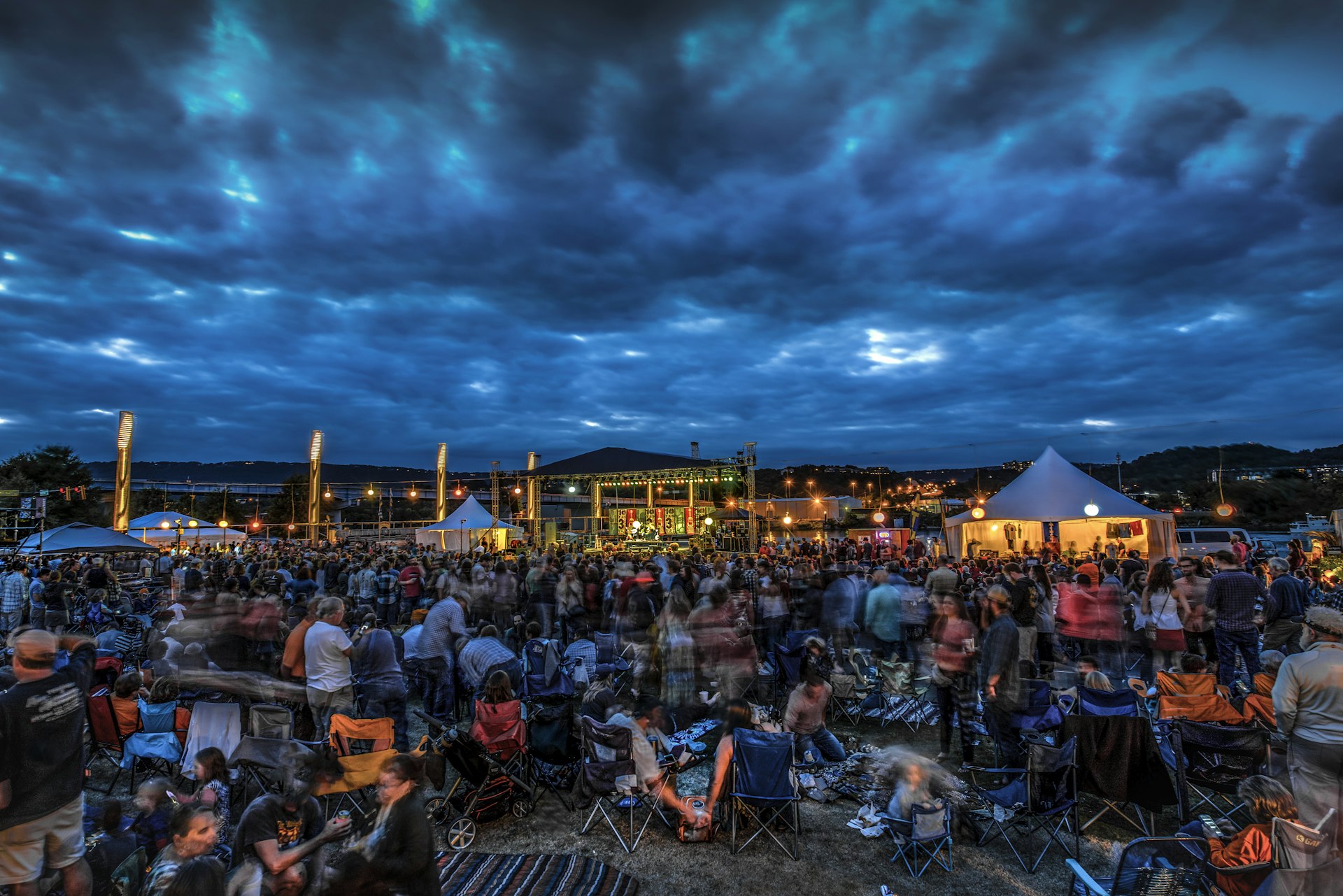 Concert crowd sat on the ground at an outdoor music festival with large dark clouds in the sky above the stage