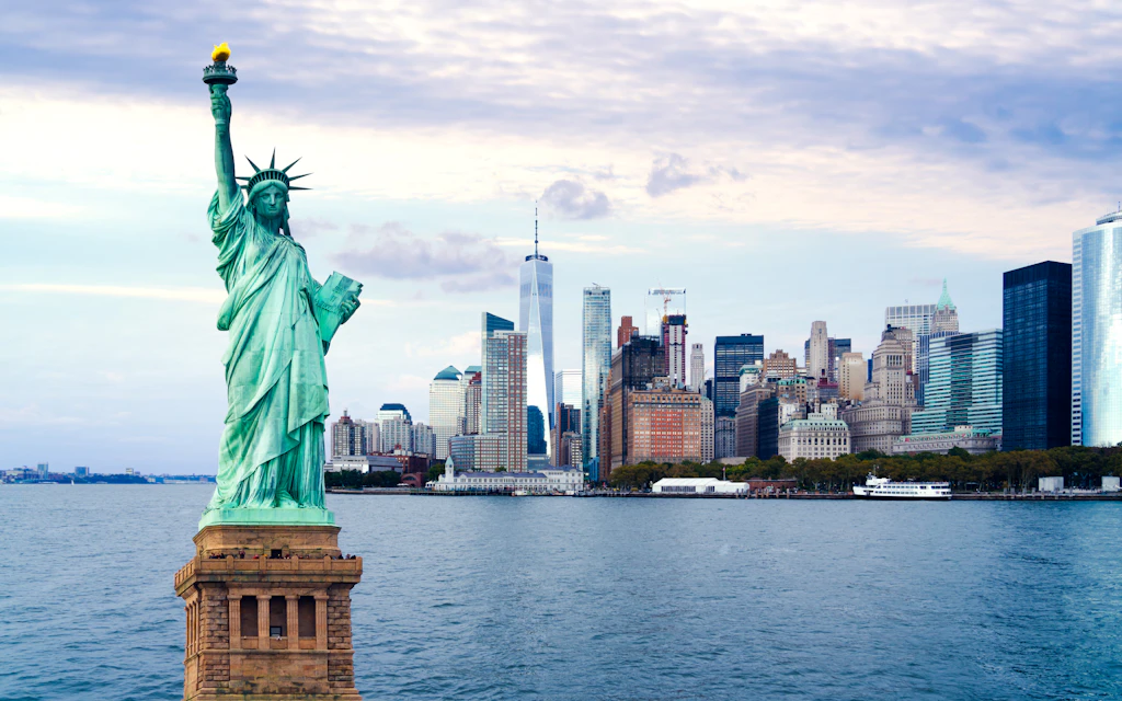 A large green copper statue of a woman with an arm in the air stands on an island in front of a cityscape of skyscrapers 