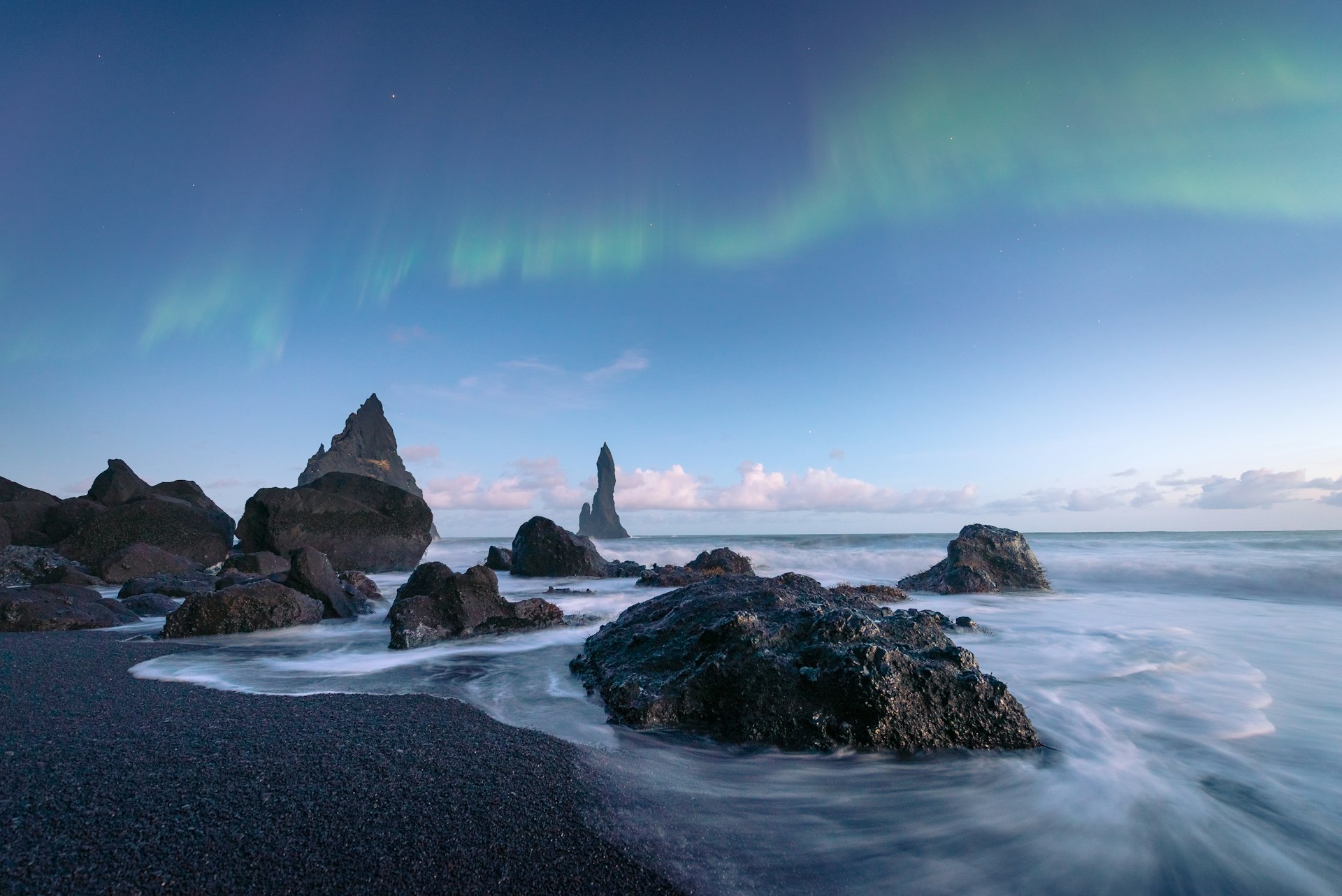 A dark-sand volcanic beach with several jagged rocky outcrops out at sea. A green streak of the northern lights dances across the sky