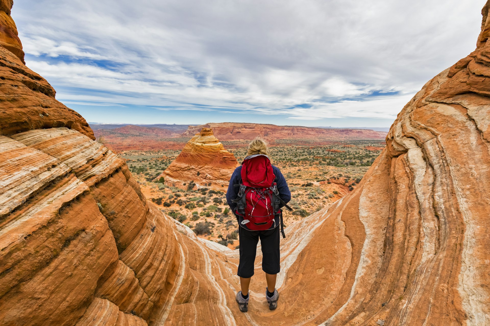 A hiker looks out to a desert landscape where the rocks are striped in orange, pink and sandstone colors