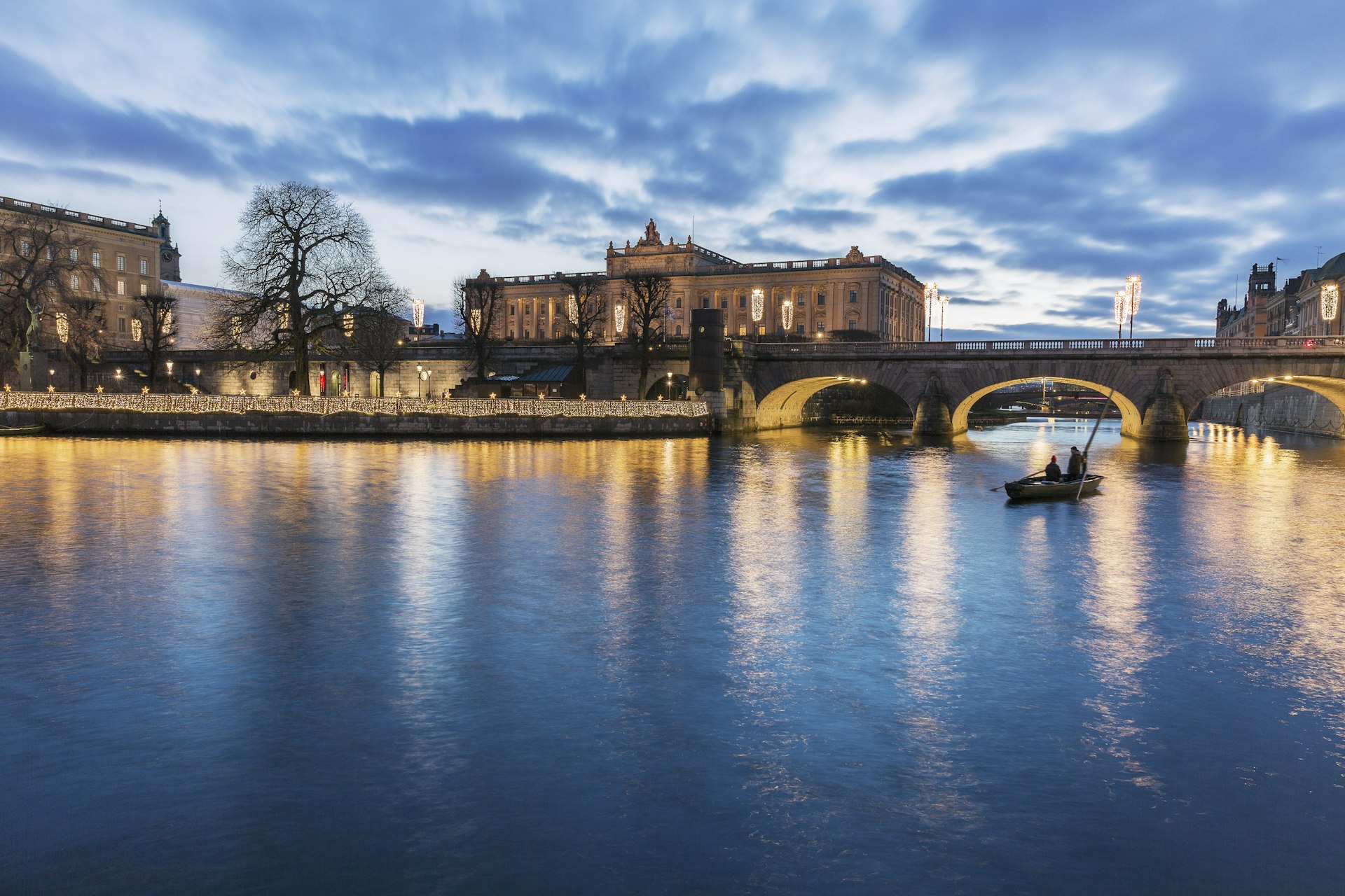 A rowboat paddles near the Norrbro Bridge in the evening with the Stockholm Palace in the background