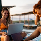 Mixed race girl and blonde girl on the beach using laptops during summer time vacation. Shot on Canon, 35mm lens iso 100. magic hour