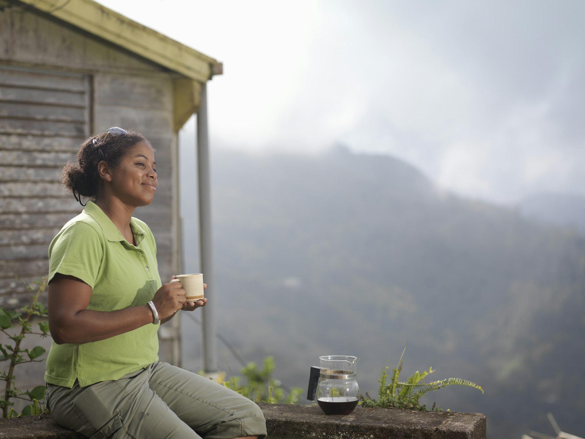 A woman wearing a green top holds a cup of coffee while looking out over the mountains in Jamaica. There is a glass coffee pot on a stone ledge.