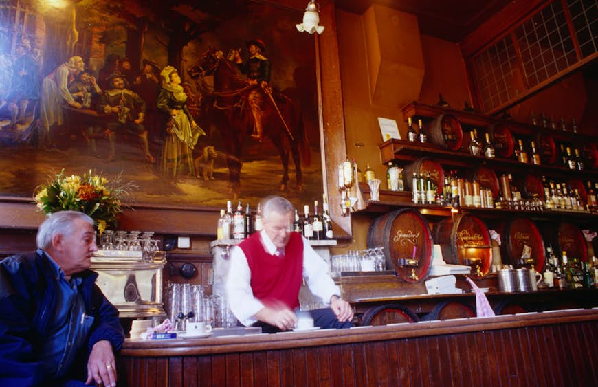 A bartender, in a dark room with heavy paintings on the walls wipes the bar, while talking to a customer