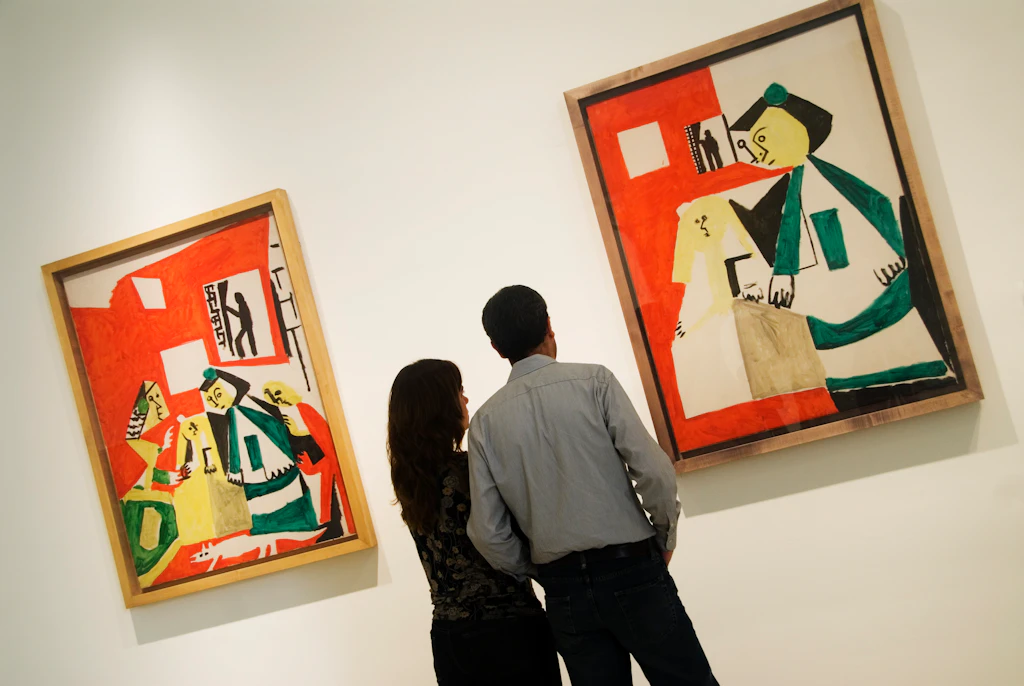 Two people stand in front of two paintings by the artist Picasso, in the Museu Picasso in Barcelona. The paintings employ Picasso's classic Cubist style.