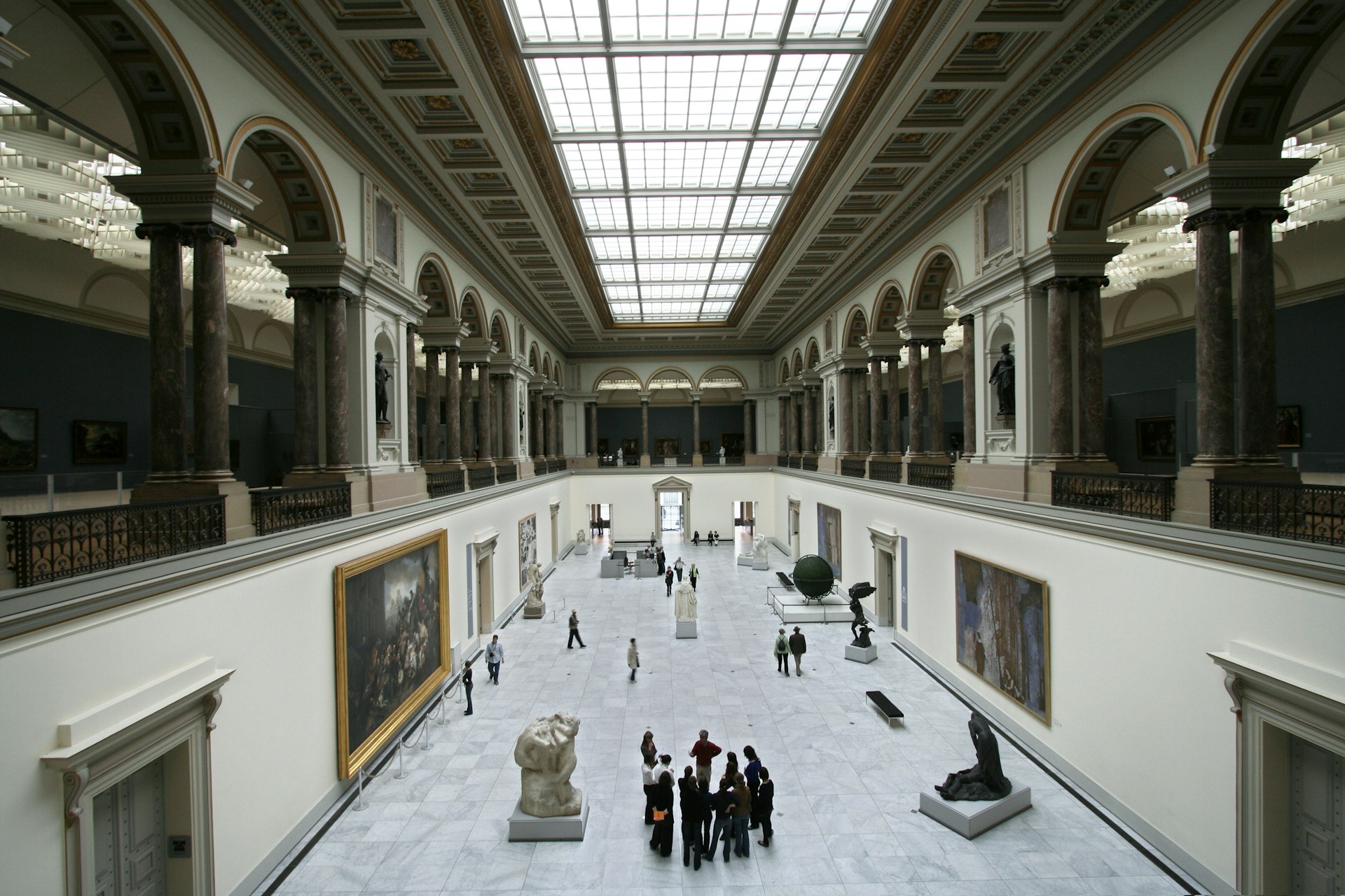 The main hall of the Musees Royaux des Beaux-Arts. The white interiors are dotted with artworks, which people walk between and stand to admire.