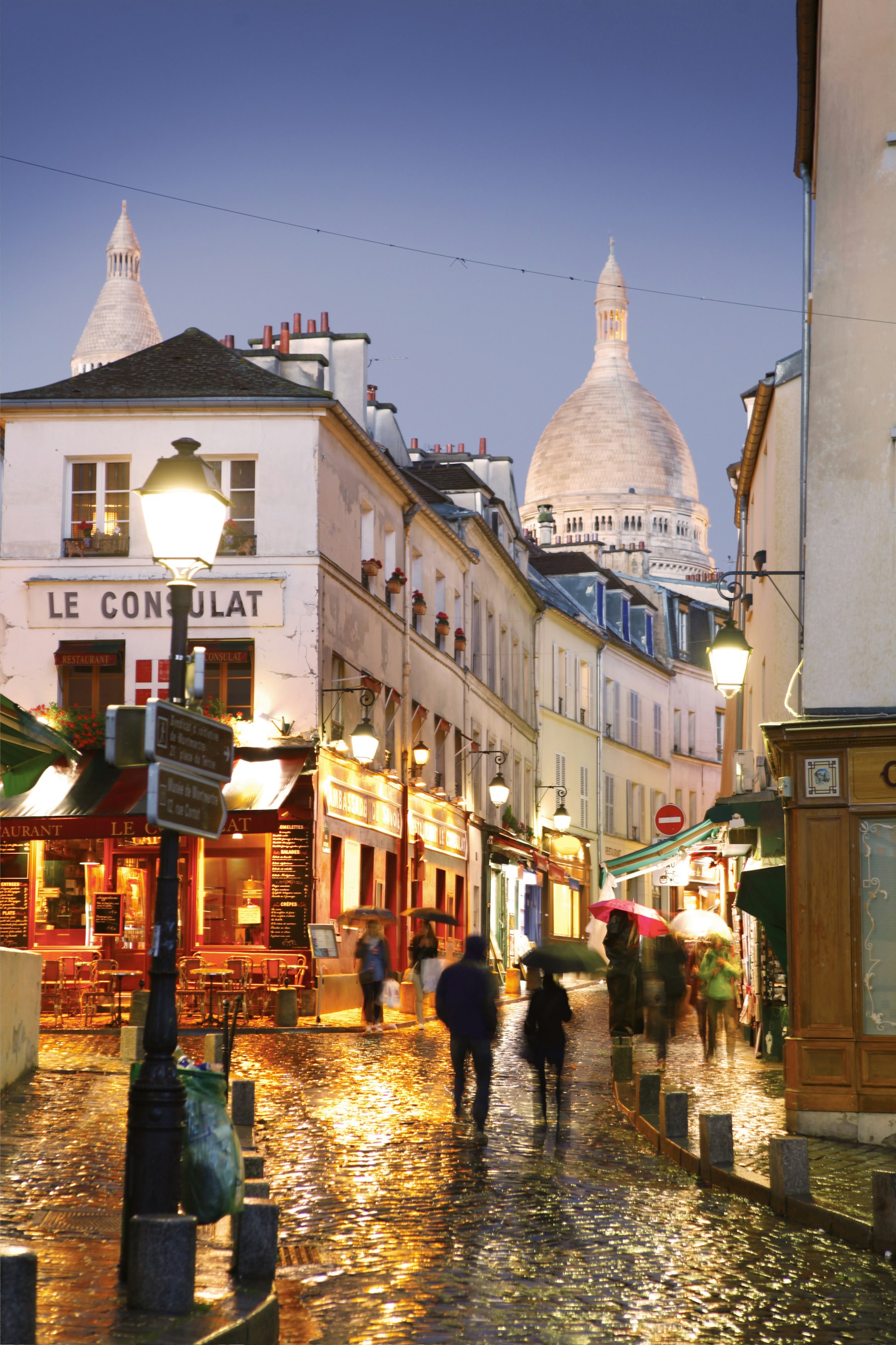 People huddled under umbrellas pass through a wet street at dusk with a huge white domed building at the top of the hill