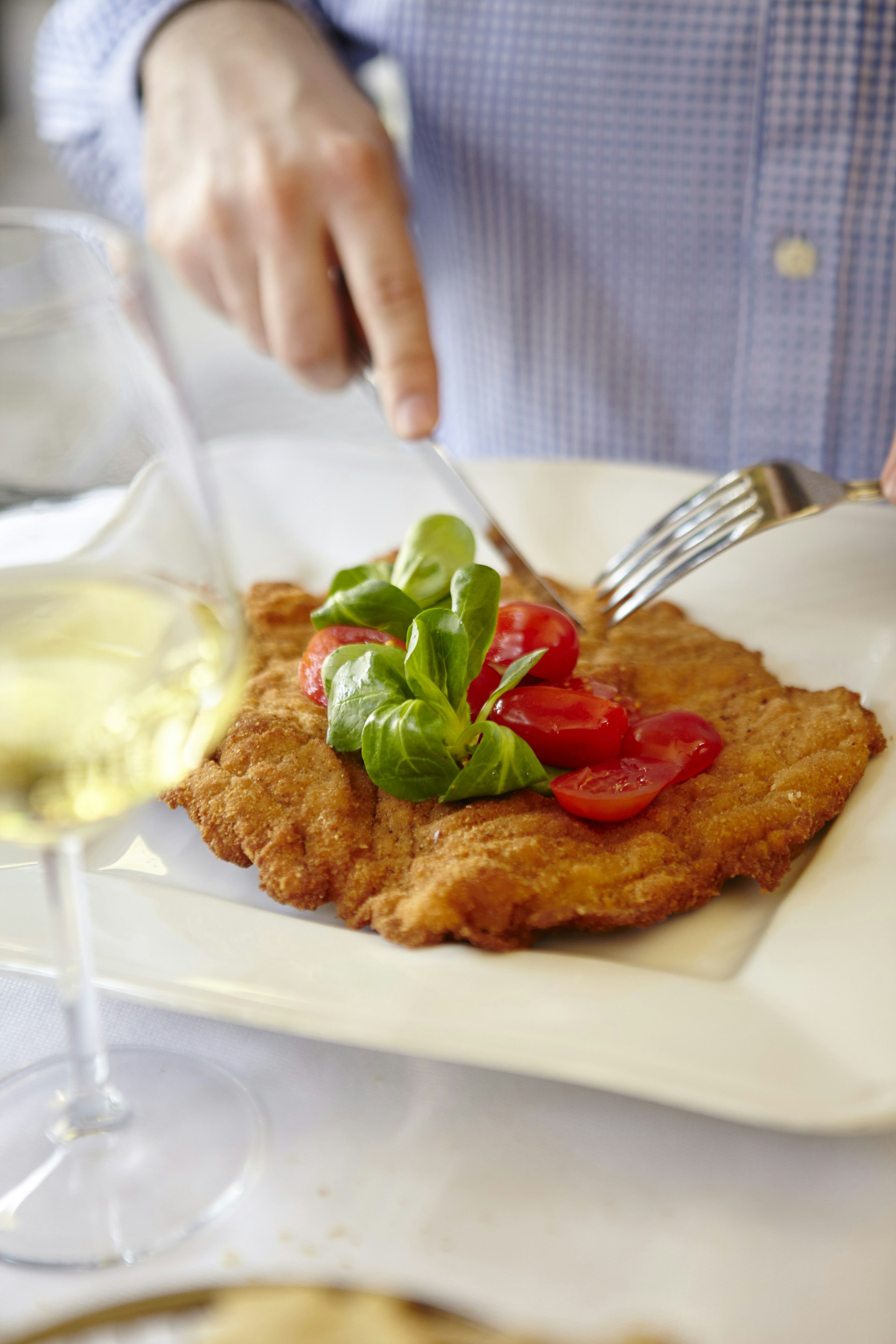 Two hands use cutlery to slice into a breaded piece of meat on a white plate