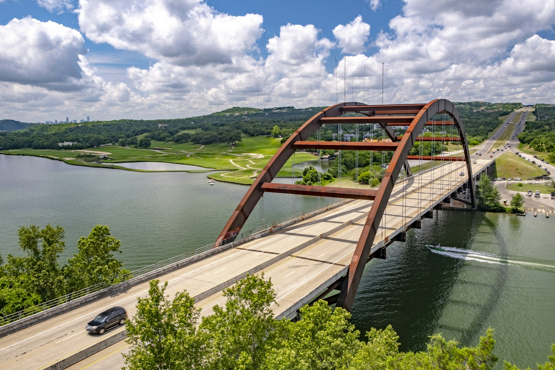 A car drives over the Pennybacker 360 bridge on the outskirts of Austin, Texas. Beyond the bridge, which spans a river, a number of Austin's skyscrapers are visible.