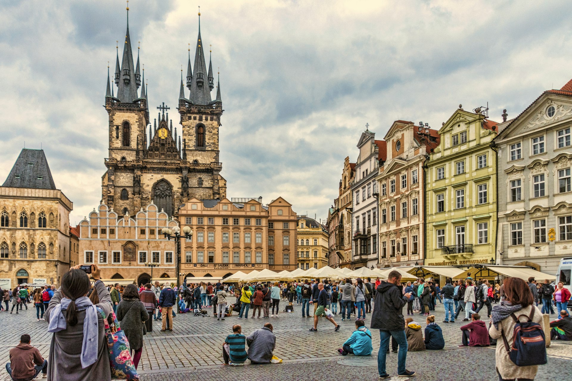 Tourists gather in a city square overlooked by twin Gothic church towers