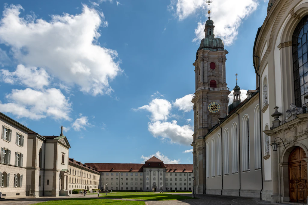 Exterior of the Abbey of St Gall. A high white wall extends into the distance, where the grand tower of a cathedral is visible.
