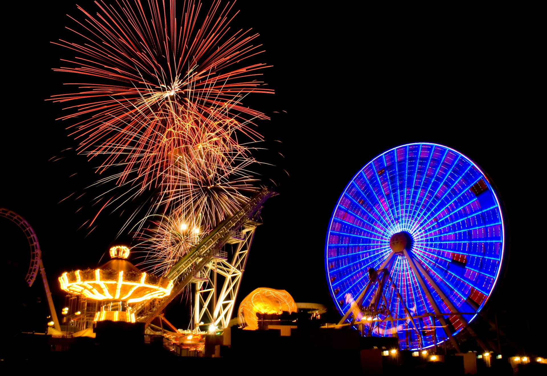 Amusement park rides, including a large ferris wheel light up at night as huge fireworks explode overhead. 