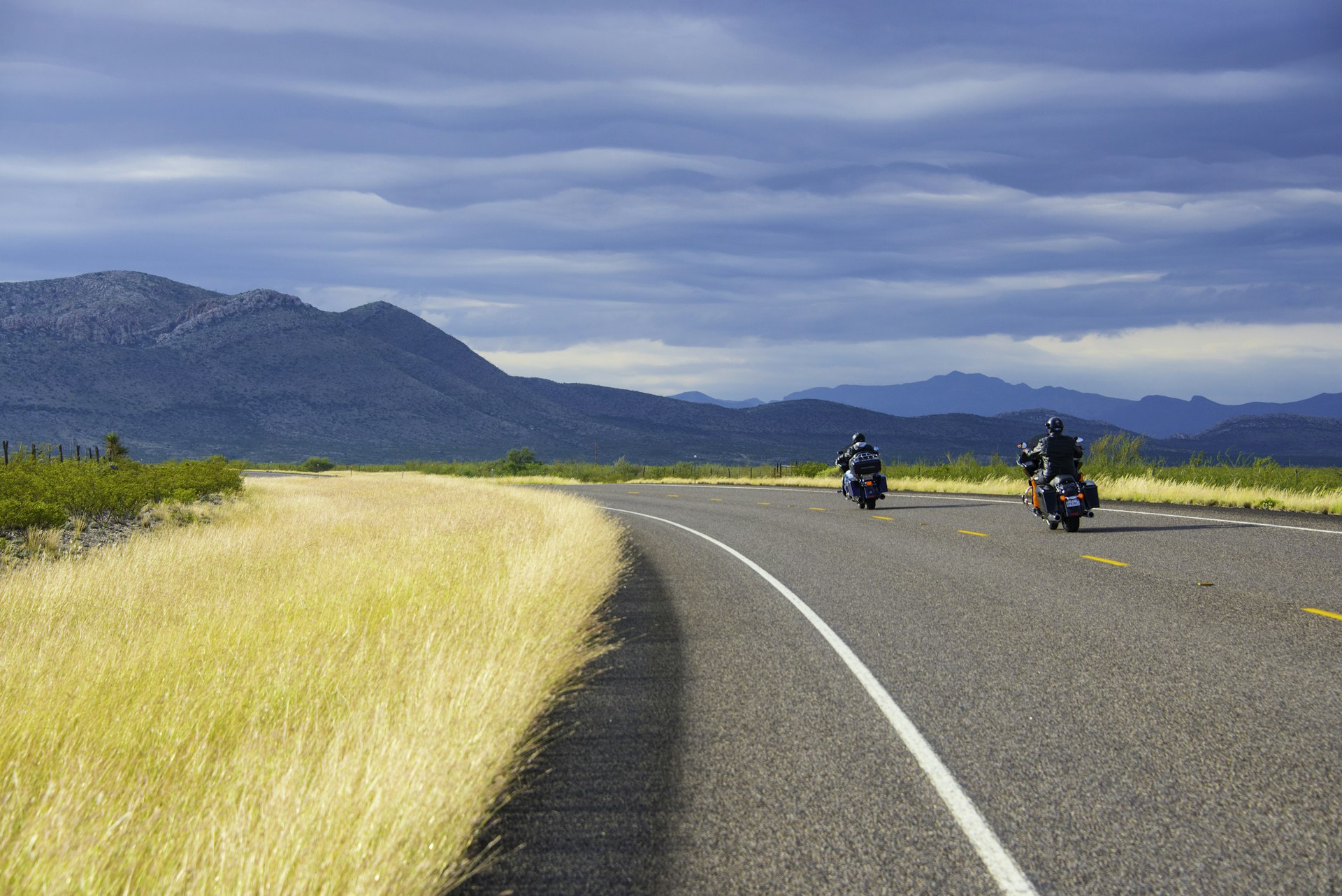 Two motorcyclists drive around a curving road in Big Bend National Park, Texas. In the distance, mountains are visible.