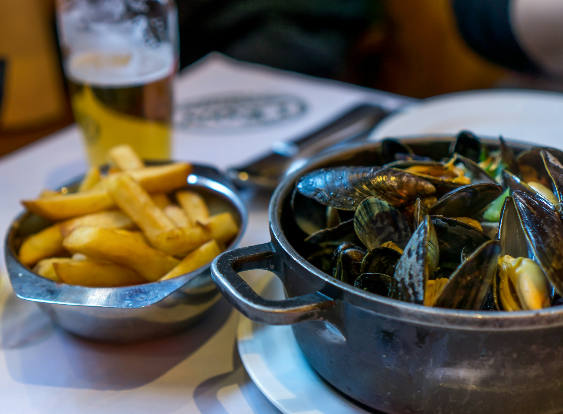 A pot of steaming mussels, with black shells and orange inners, stand on a table next to a plate of fries and a pint of beer.