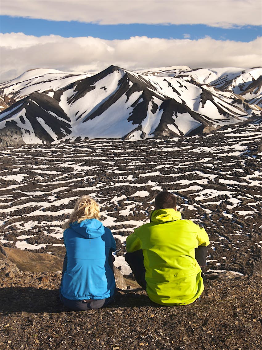 Two people sit with their backs to the camera looking out over a black lava field and mountainous landscape
