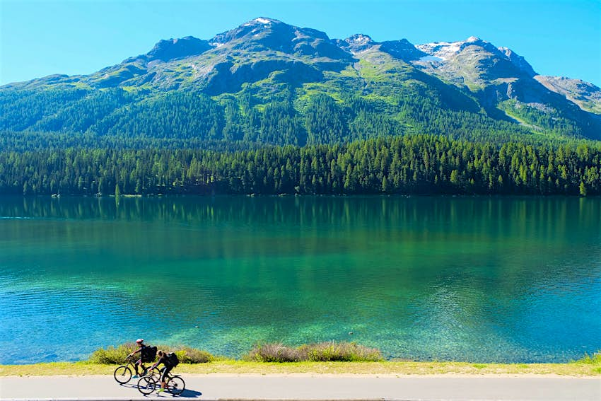 Radfahrer entlang einer Uferstraße in Lugano, Schweiz.  Hinter dem großen See sind Berge zu sehen.