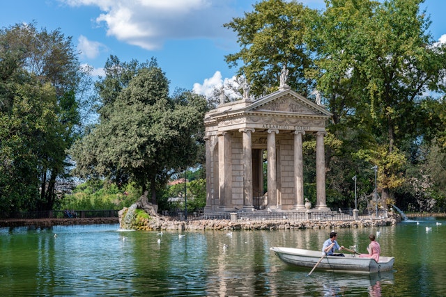 Two people in a row boat with a temple-like building in the center of the lake