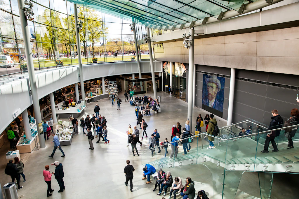 A view of the interior of the Vincent Van Gogh Museum in Amsterdam. The museum is filled with people, who walk between exhibits. The museum is very light, with many windows and walls made of glass.