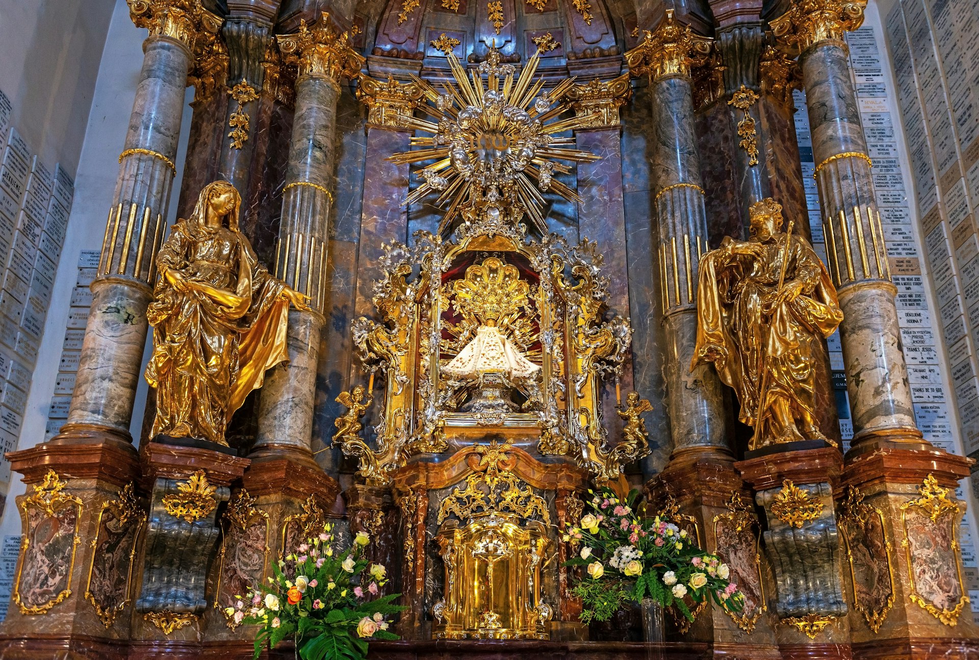 A small statue of an infant Jesus stands in the centre of a lavish, golden shrine in a church in Prague. The statue is wearing a white cloak.