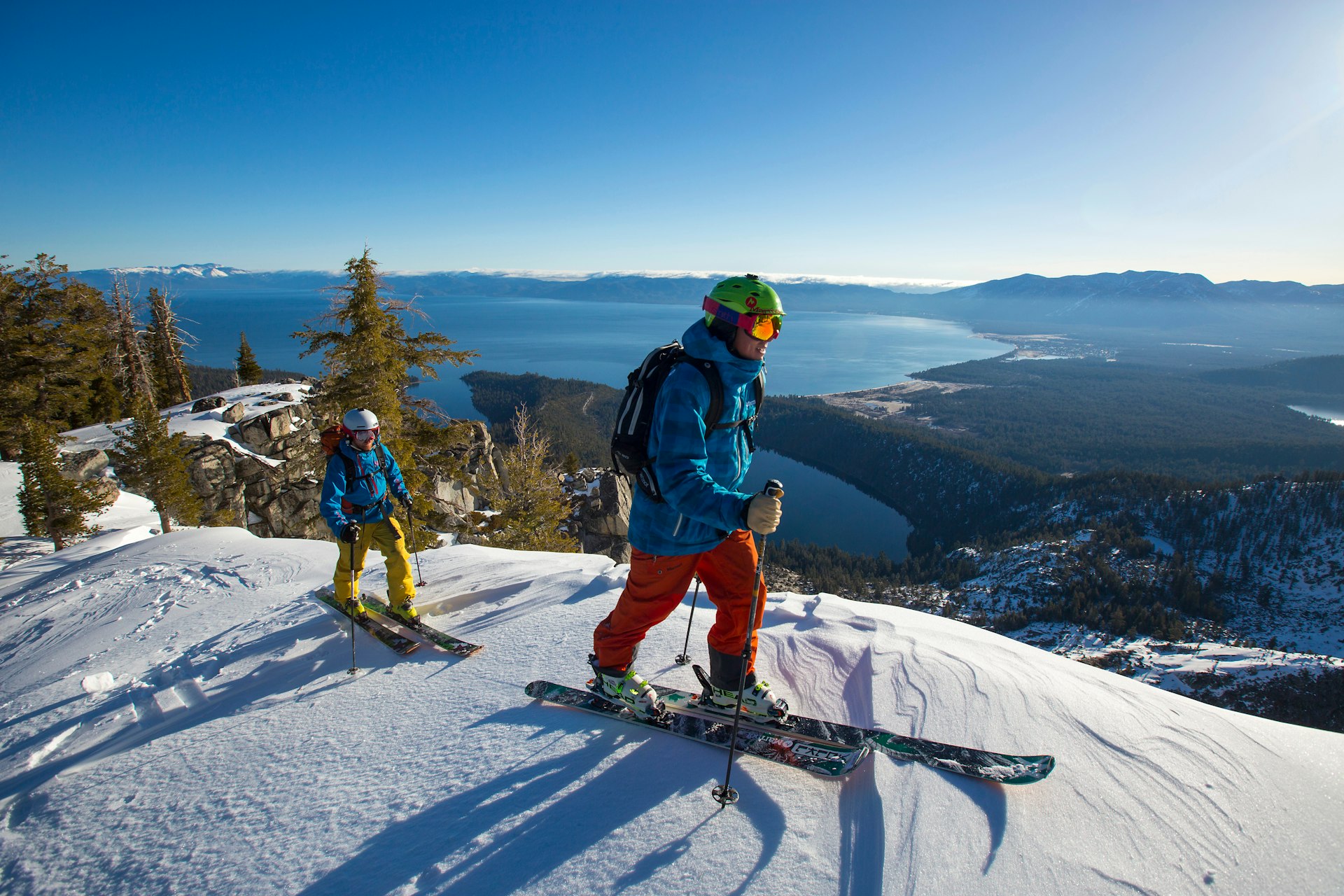 An adult and a child in full snow gear cross-country-ski along a snowy ridge high above a lake