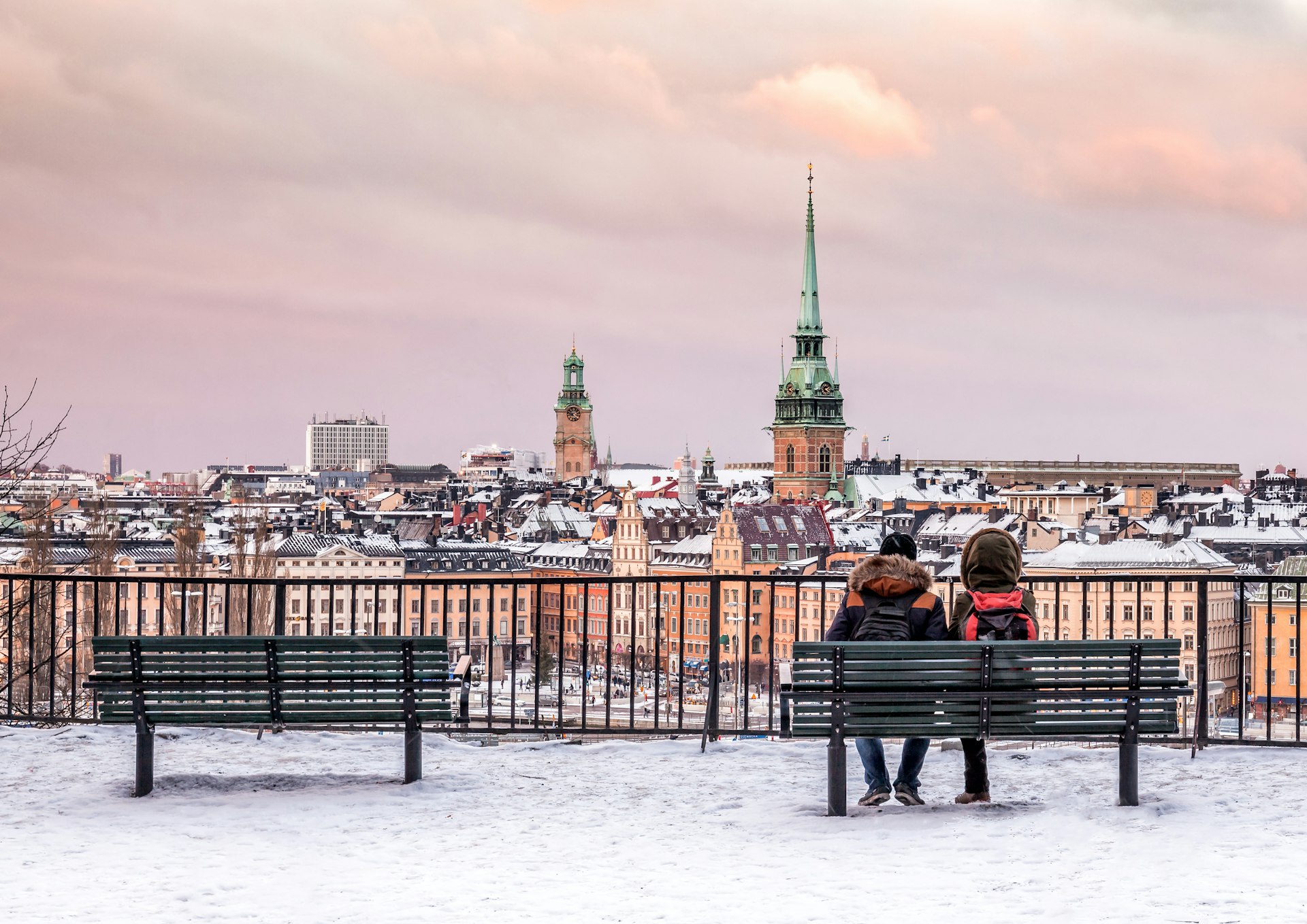 A couple sit at a bench with their backs to the camera looking towards the snow-covered roofs and spires of an Old Town city center