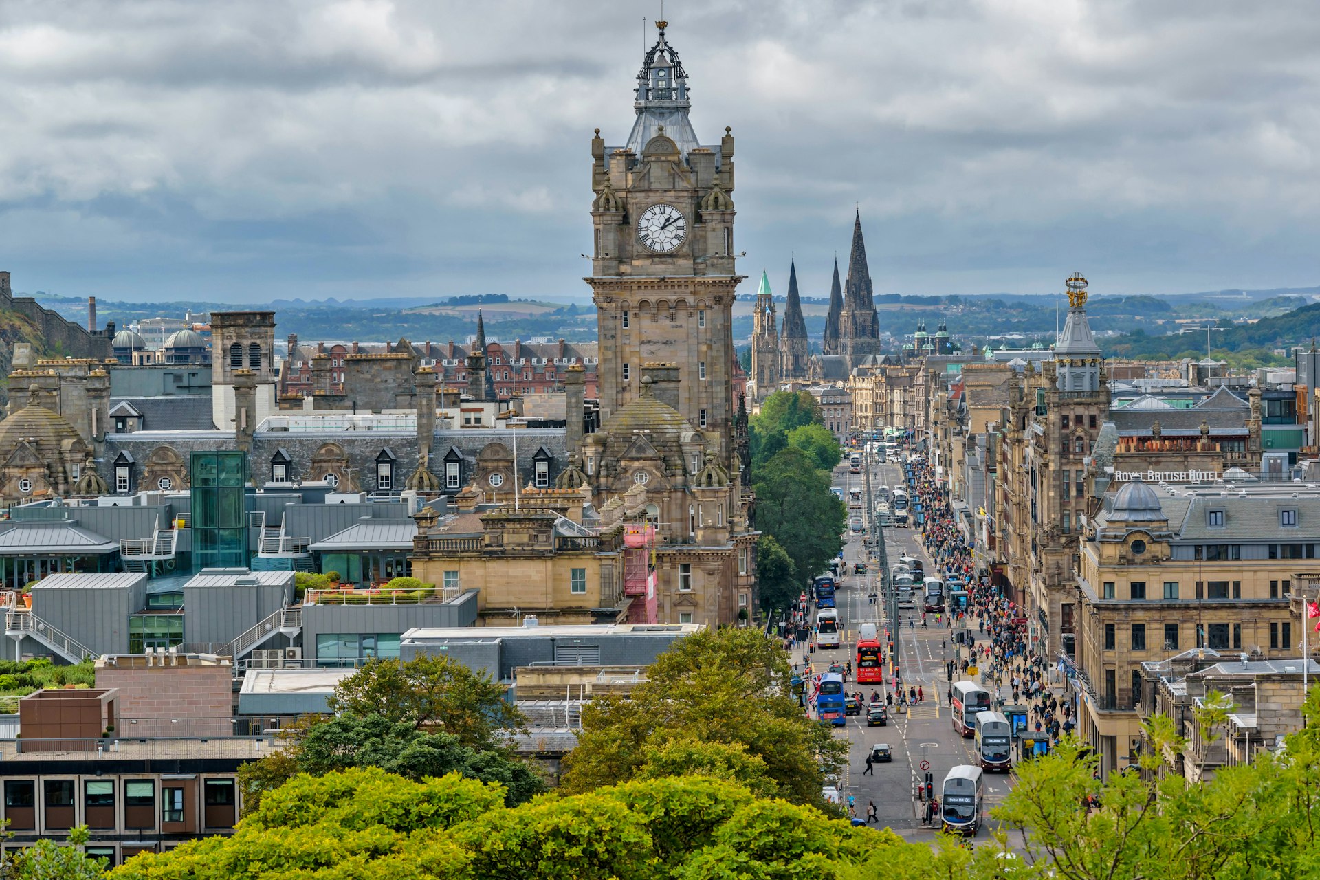 The Balmoral Hotel clock tower stands above on Princess street in Edinburgh with a queue of traffic, including a red bus, making its way along the street to the green hills in the background