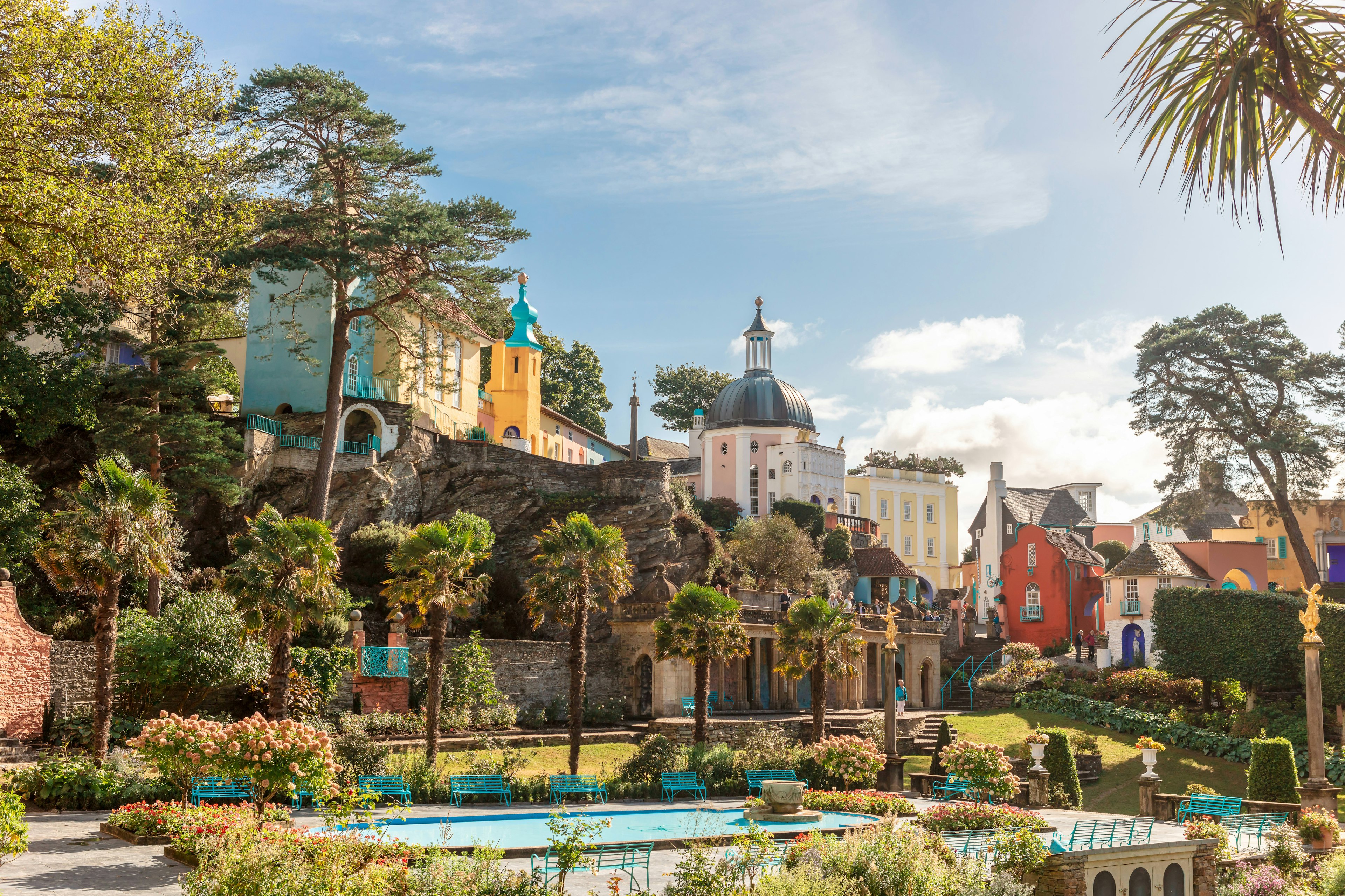 Italian-style buildings in pastel colors with palm trees and benches surrounding a large rectangular water feature