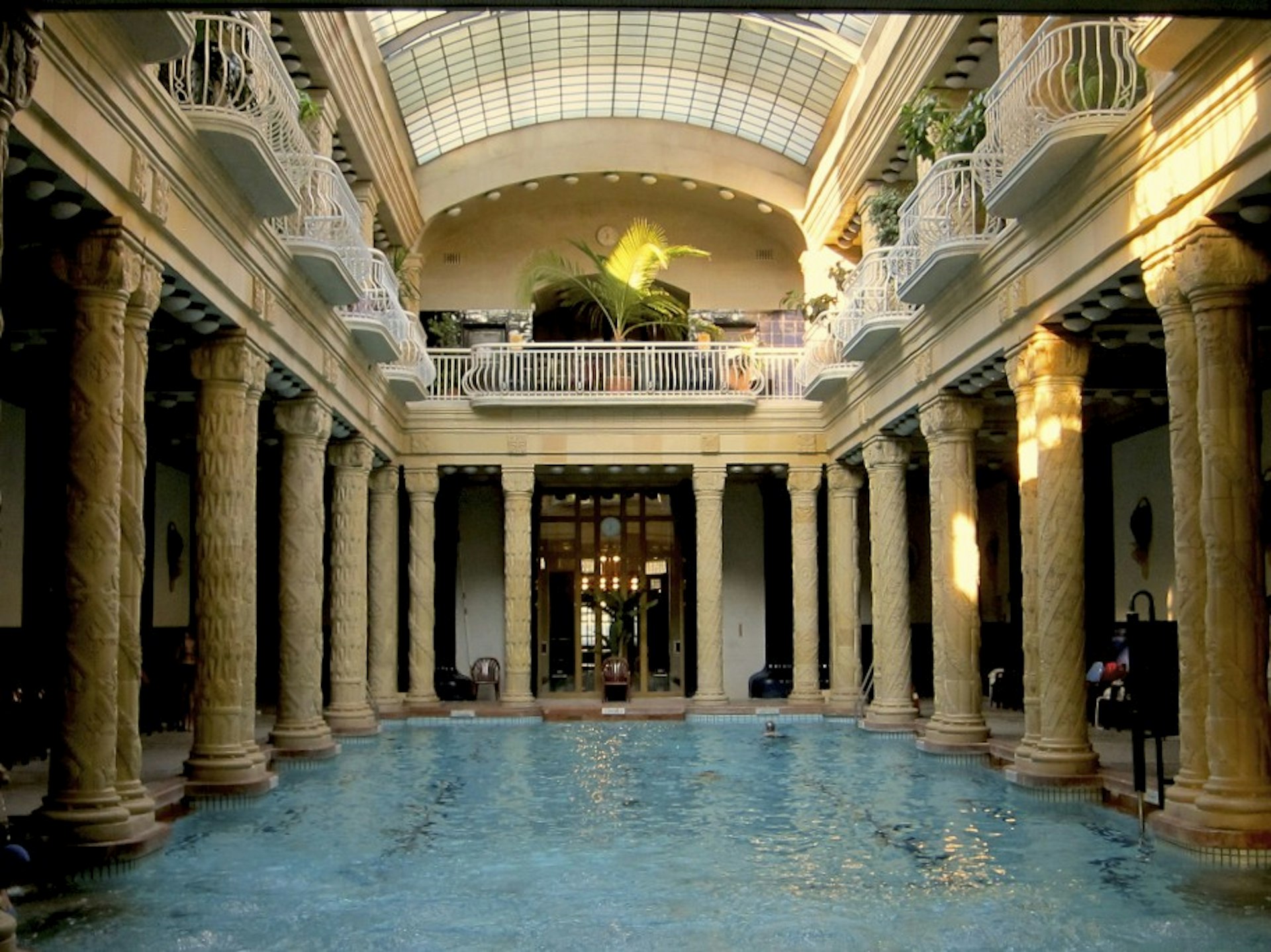 The cram-colonnaded indoor pool at Gellért Baths, Budapest shimmers in the sunlight that lands on its surface through the glass-roof like crepuscular rays on the ocean. The pool is surrounded one floor up by white Juliet balconies. 