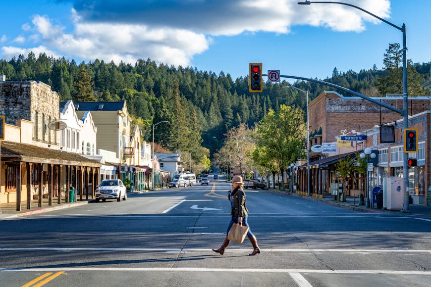 A woman in a hat, jacket, and boots crosses the street in quaint downtown Calistoga, California, with tall pines in the background
