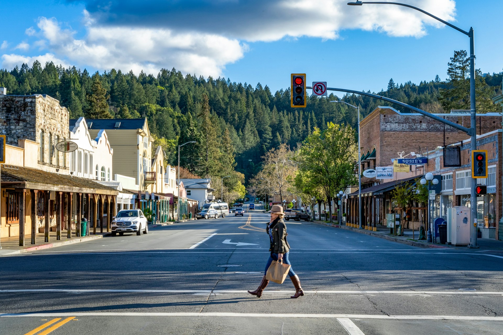 A woman in a hat, jacket, and boots crosses the street in quaint downtown Calistoga, California, with tall pines in the background
