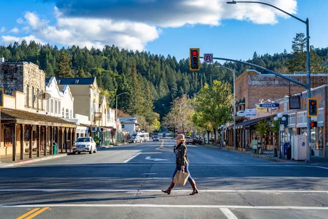 A woman in a hat, jacket, and boots crosses the street in quaint downtown Calistoga, California, with tall pines in the background