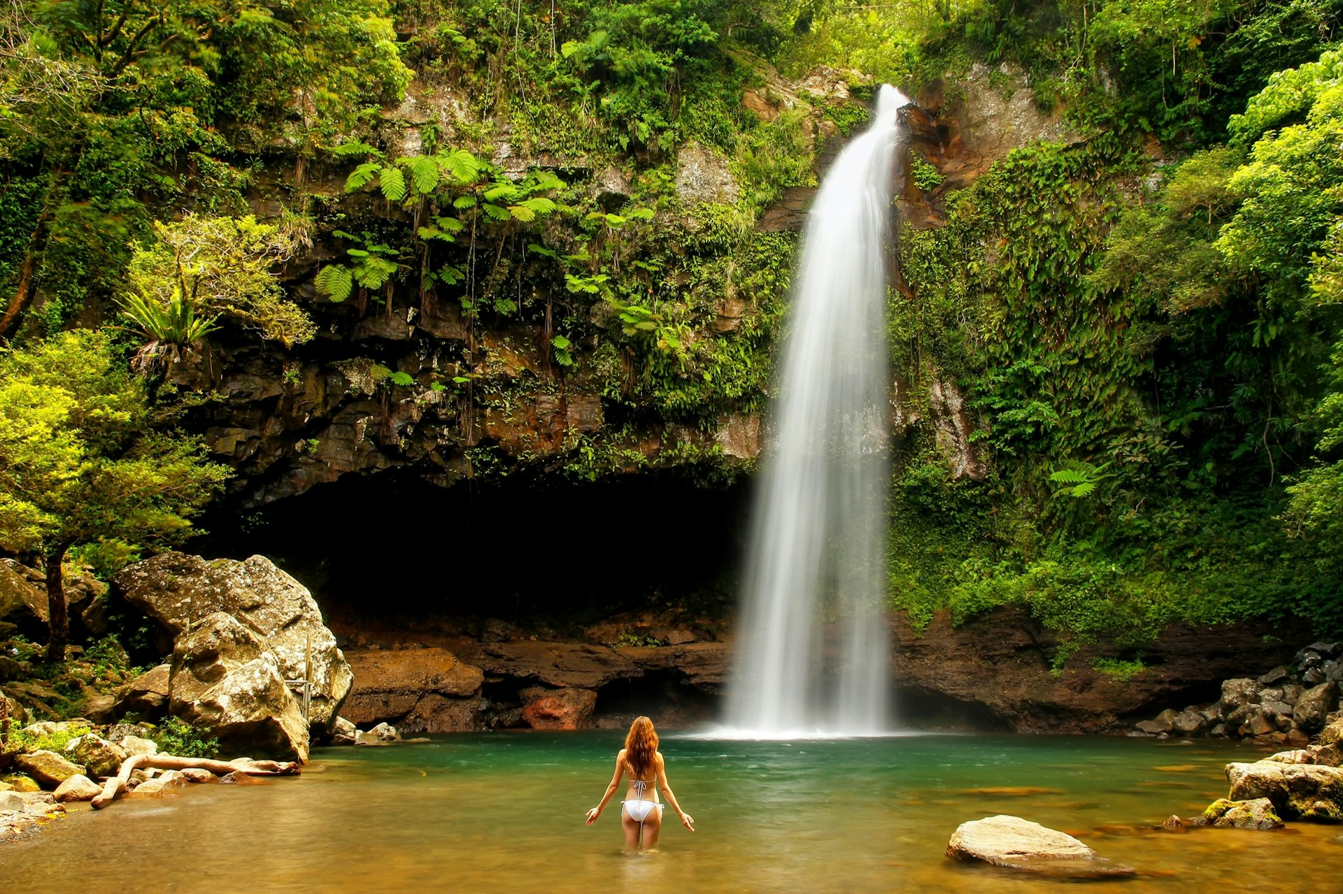 A woman stands in the water facing a tall waterfall surrounded by lush foliage