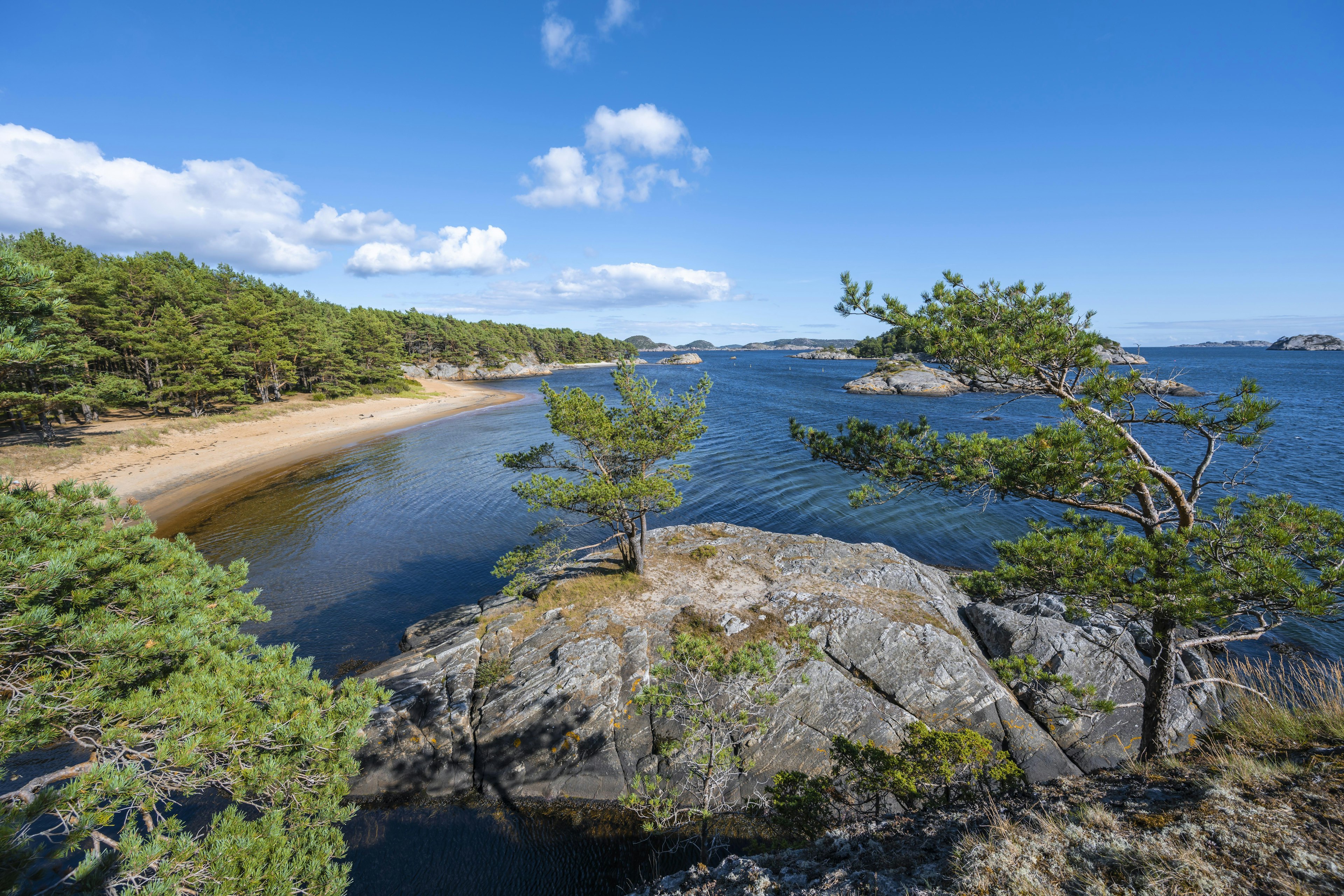 A view of Furulunden nature park, with two sandy coves visible; both of which are lapped by dark blue seas. The hillsides are bright green, filled with plants and trees.