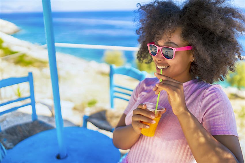 Greek Islands A woman sitting by the sea smiling and drinking a glass of juice