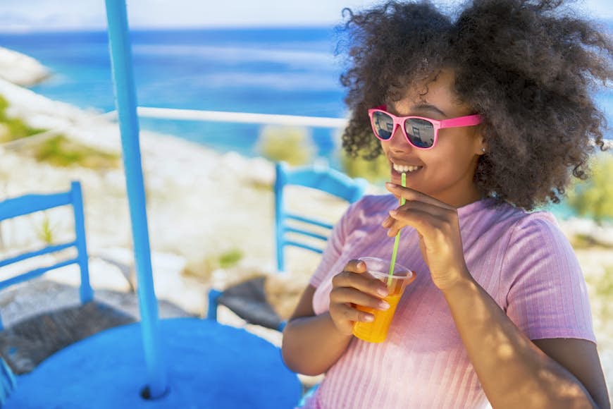 A woman is sitting in the sunshine smiling and drinking from a glass