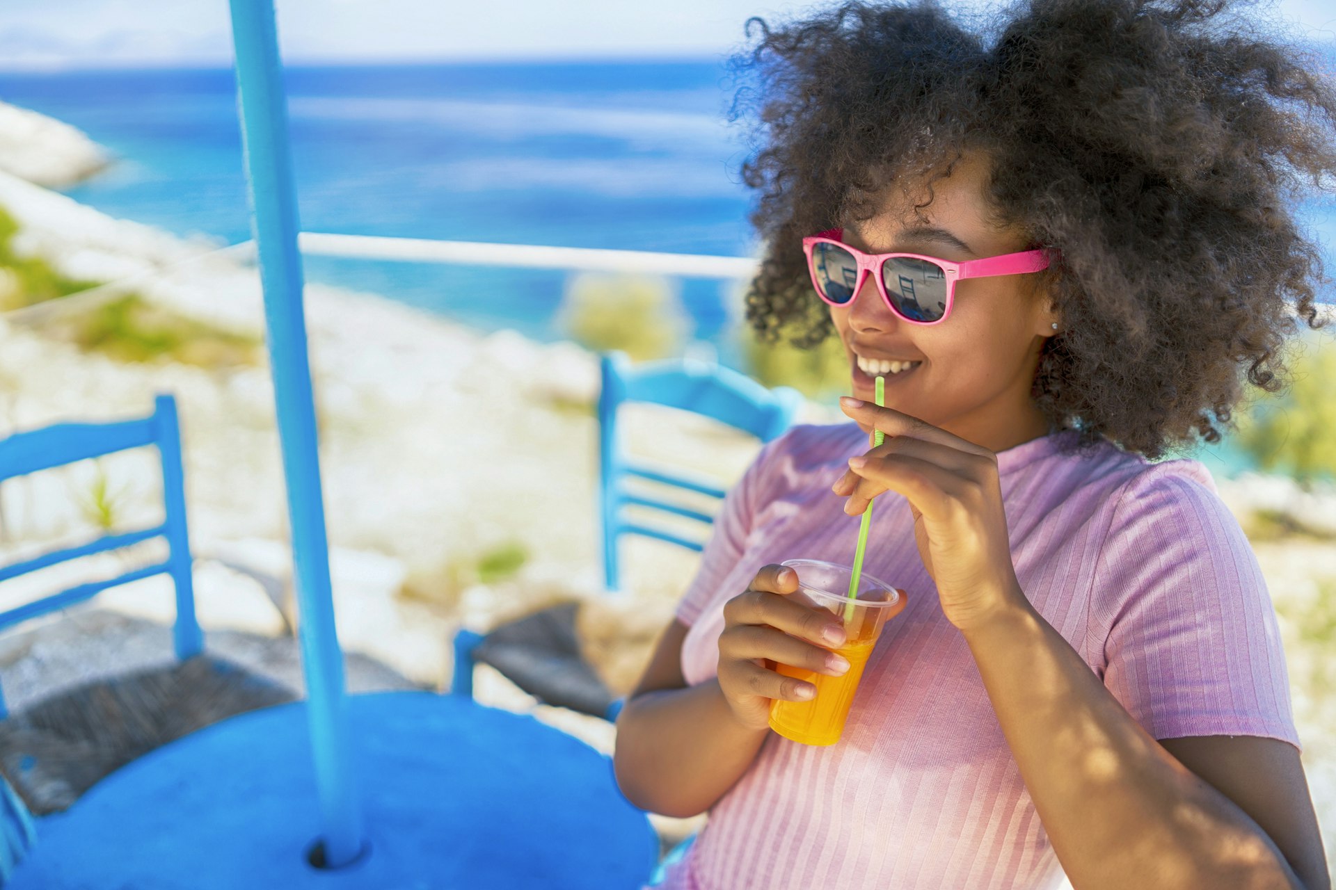A woman sitting by the sea smiling and drinking a glass of juice