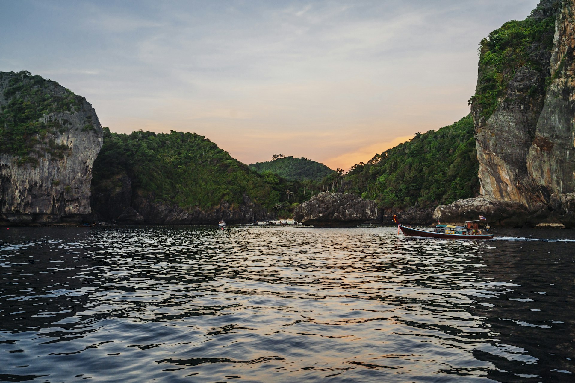 A shot taken from the water facing towards the shore, which is dotted with large rocky cliffs covered in vegetation. A small colorful fishing boat sails by