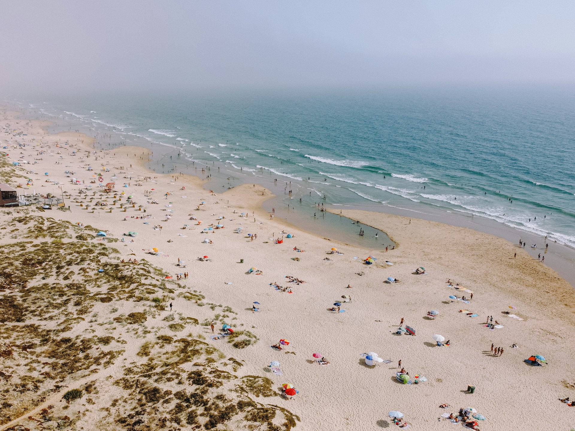 An aerial shot of a large sweep of sandy beach with many groups of people sat on the sand under colorful umbrellas